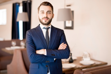 Serious ambitious bearded young restaurant manager in formal suit crossing arms on chest and looking at camera in cozy public establishment